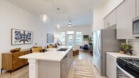 This thoughtfully designed 1-bedroom, 1-bathroom residence offers a contemporary open-concept layout bathed in natural light. Light wood-tone flooring runs throughout, tying together each space seamlessly. The kitchen serves as the heart of the home, featuring a central island with a white quartz countertop, undermount sink, pendant globe lighting, gray shaker cabinetry, a subway tile backsplash, and stainless steel appliances including a gas range, built-in microwave, dishwasher, and French door refrigerator. The kitchen flows directly into the living area, which is anchored by a ceiling fan and large windows and a glass-panel door that invite outdoor views and generous natural light. The bedroom is a serene retreat with multiple windows framed by elegant draping curtains, providing ample daylight and pleasant exterior views. The bathroom offers a clean, modern aesthetic with gray cabinetry, a white quartz vanity, large-format tile flooring, and direct access to a spacious walk-in closet equipped with dual hanging rods and built-in shelving. The entrance completes this efficiently organized and stylishly finished residence.
