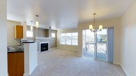 This well-appointed single-floor residence offers an open-concept layout that seamlessly connects the living and kitchen areas, creating an inviting and functional environment. The living area is anchored by a sleek, built-in fireplace framed in crisp white, providing a warm focal point for the room. Plush, neutral-toned carpeting extends throughout, adding comfort underfoot and complementing the warm beige walls. Natural light pours in through large windows that frame views of the surrounding neighborhood, enhancing the bright and airy atmosphere. A set of glass doors opens directly onto a private balcony, extending the living space outdoors and offering open urban views — perfect for relaxing or entertaining. The kitchen area features a raised breakfast bar that partially separates it from the living space while maintaining the open feel. Elegant pendant lighting is thoughtfully placed in both the kitchen and dining areas, adding a touch of style and warmth. A multi-light chandelier near the balcony entrance further elevates the interior's sophisticated character. The overall ambiance is clean, modern, and welcoming, with a cohesive neutral palette that provides a versatile backdrop for a variety of interior styles.