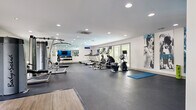 The Highland Clubhouse is a well-appointed single-story community facility designed to serve as a central gathering and amenity hub. The layout flows seamlessly from zone to zone, with wood-look flooring, recessed lighting, and neutral-toned walls creating a cohesive, modern aesthetic throughout.

At one end of the building sits a fully equipped fitness center featuring cardio machines, a cable weight system, free weights, and floor space for stretching and mat exercises. Large windows and wall mirrors enhance the sense of openness, while motivational wall art adds energy to the space.

The central section of the clubhouse includes a full kitchen outfitted with dark cabinetry, granite countertops, stainless steel appliances, and a large island with bar seating. Sliding glass doors lead to an outdoor view, allowing natural light to fill the area. Adjacent to the kitchen is a casual dining nook with round tables and upholstered chairs positioned near windows with slatted blinds.

The lounge area features comfortable seating arrangements anchored by a patterned area rug, a wall-mounted television, and decorative accent mirrors. A dedicated mailroom with a full bank of individual mailboxes connects directly to the lounge, adding everyday convenience. The overall ambiance is clean, functional, and inviting.