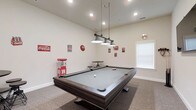 This single-floor space is designed as a dedicated entertainment and game room, offering a lively yet comfortable atmosphere for recreation and socializing. The centerpiece of the room is a full-size billiards table finished in dark wood with a light gray felt surface, positioned beneath a stylish triple pendant light fixture with industrial-style metal shades that provide focused overhead illumination. The room features neutral-toned walls and carpeted flooring that contribute to a relaxed, casual ambiance. Recessed lighting throughout the ceiling supplements the pendant fixture, ensuring the space is well-lit from multiple angles. A charming retro aesthetic is established through a collection of vintage-style Coca-Cola signs and metal wall decorations, adding personality and visual interest to the room. A classic red popcorn machine stands in the corner, reinforcing the entertainment theme and serving as both a functional and decorative element. A small bistro-style table and seating are visible in the background, providing a spot to relax between games. A window with white blinds allows natural light to filter into the space. The room's generous proportions and thoughtful layout make it an ideal setting for hosting gatherings and enjoying leisurely activities.
