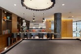 Located at 4200 N Carlin Springs Rd in Arlington, VA, this thoughtfully designed amenity lounge offers a sophisticated and multi-functional gathering space across a single floor. The space is anchored by striking oxidized cylindrical columns that lend an industrial-chic character throughout. Wide-plank hardwood flooring flows through the open layout, complementing the warm tones of the wood-paneled walls and built-in shelving.

A full billiards table takes center stage in the main area, while a dark granite-topped bar with leather upholstered barstools serves as a social hub, illuminated by elegant pendant lighting with sculptural designs. Adjacent to the bar, a lounge nook features built-in bookshelves lined with curated volumes and decorative objects, paired with plush seating and an artfully patterned area rug.

Vibrant rust-orange lounge chairs clustered around a glass-top table create an inviting conversation area near a glass-railed mezzanine overlooking a double-height atrium. The atrium showcases a dramatic cascading globe chandelier framed by floor-to-ceiling windows that flood the space with natural light and exterior views. A wood-paneled elevator bay adds warmth and texture, while curated artwork and sculptural accents throughout reinforce the space's refined, contemporary aesthetic.