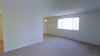 This single-floor space offers a clean, functional layout characterized by a neutral color palette and an open, airy feel. The interior is finished with wall-to-wall plush carpeting in a warm beige tone, providing a soft and comfortable underfoot experience throughout. The main living area is generously proportioned, allowing for flexible furniture arrangements to suit a variety of needs.

A large window with vertical blinds brings in an abundance of natural light, brightening the space and creating a welcoming ambiance. The smooth, white walls contrast gently with the textured ceiling, adding subtle visual depth without overwhelming the simplicity of the design.

A pendant light fixture is positioned in an adjacent area, suggesting a defined dining or secondary living zone just off the main room. The transition between these areas is marked by a partial wall, which provides a sense of delineation while maintaining an open and connected feel.

Overall, the space presents a straightforward, well-maintained interior that benefits from its bright, open character and clean finishes. The understated design elements make it a versatile canvas, well-suited to a range of personal styles and furnishing preferences.