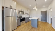 This single-floor residence offers a clean, contemporary design highlighted by warm wood-tone luxury vinyl plank flooring that flows consistently throughout the main living areas. The kitchen serves as the centerpiece of the space, showcasing a two-tone cabinetry scheme that pairs crisp white upper cabinets with darker gray lower cabinets, all accented by sleek hardware. A classic white subway tile backsplash runs along the cooking wall, complementing the bright white countertops. A full suite of stainless steel appliances — including a refrigerator, electric range with over-the-range microwave, and dishwasher — equips the kitchen for everyday use. A center island provides additional prep space and storage, illuminated overhead by a pair of stylish glass pendant lights. Recessed lighting throughout ensures a well-lit environment. The open living area connects seamlessly to a private balcony through a large sliding glass door, allowing natural light to fill the space and offering pleasant outdoor views of surrounding greenery. A carpeted room adjacent to the living area provides a softer, quieter retreat. The bathroom features wood-look flooring, a dark vanity, and grab bars, reflecting a practical and accessible design. Neutral walls throughout keep the aesthetic versatile and inviting.