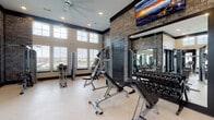 Located at 860 Candlelite Ln in Lewis Center, Ohio, this well-appointed fitness center delivers a comprehensive workout experience within a thoughtfully designed, single-floor facility. The space is defined by soaring double-height ceilings and expansive windows that flood the interior with natural light, creating an open and energizing atmosphere throughout.

The cardio zone is equipped with multiple treadmills, a rowing machine, stationary bikes, and an elliptical trainer, all positioned to take advantage of the bright, airy environment. A dedicated strength training area offers a full rack of dumbbells, adjustable benches, and a cable machine system, complemented by large wall-mounted mirrors that enhance the sense of space. A mounted flat-screen television is positioned above the strength area for added convenience.

The interior design blends modern and industrial aesthetics, featuring a striking exposed brick accent wall, bold dark trim work, light herringbone-patterned flooring, and recessed lighting throughout. Black-framed French doors connect the fitness area to an adjacent space, adding architectural depth.

Convenience amenities include a dual water fountain and bottle-filling station set against a decorative geometric tile wall, as well as easily accessible restrooms. The overall ambiance is clean, polished, and motivating — a high-quality fitness facility suited for a wide range of workout routines.