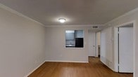 This single-floor space offers a clean, functional layout designed for comfortable everyday living. The main living area features warm hardwood-style flooring, crisp white walls, and elegant crown molding that adds a classic architectural touch. A textured ceiling with a centrally mounted light fixture provides even illumination throughout the room, while a ceiling vent ensures efficient climate control.

A distinctive pass-through window connects the living area to the kitchen, creating an open, communicative feel between the two spaces. The kitchen is equipped with gray cabinetry, a stainless steel sink, and a black refrigerator, offering a modern, understated aesthetic. The cabinetry provides ample storage, and the layout appears well-organized for practical use.

A doorway visible from the living area leads to additional rooms, suggesting a connected and accessible floor plan. Baseboards are cleanly finished throughout, complementing the overall neutral color palette of gray and white tones. The space is straightforward in its design — prioritizing utility and ease of movement — making it well-suited for those seeking a low-maintenance, single-level living environment with cohesive finishes and a tidy, modern appearance.