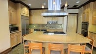 This single-floor space centers around a thoughtfully designed kitchen that balances professional functionality with an inviting atmosphere. The focal point is a generously sized, curved granite island topped with warm golden stone, surrounded by wood-framed bar seating — ideal for casual dining or social gatherings during meal preparation.

The kitchen is equipped with high-end, professional-grade appliances, including a large stainless steel built-in refrigerator and a multi-burner gas cooktop integrated into the island, topped by a substantial stainless steel range hood that anchors the space visually. A secondary prep area along the back wall features a stainless steel sink, additional cabinetry, and built-in appliances beneath a decorative tile backsplash.

Cabinetry throughout is crafted in a light natural wood tone with clean lines, extending to the upper walls and complemented by dark crown molding that adds a refined, finished look. Storage is generous, with tall pantry-style cabinets featuring distinctive X-panel detailing adding architectural character.

Overhead, a combination of recessed lighting and warm pendant fixtures provides layered illumination. Sheer drapery panels add a soft, elegant touch, enhancing the overall ambiance of this well-equipped and welcoming kitchen space.