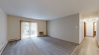 This single-floor residence features four bedrooms and two bathrooms, thoughtfully arranged within a compact and efficient floor plan. The living room is a standout space, featuring plush grey carpeting, a sliding glass door with vertical blinds that invites natural light and provides outdoor views, and a baseboard heating unit along the wall. The open feel of the room makes it ideal for everyday living and entertaining. The kitchen is well-appointed with bold dark cabinetry, light countertops, and white appliances including a refrigerator and electric range. Light wood-style flooring runs throughout the kitchen and connects seamlessly to the adjacent dining area. Each of the four bedrooms offers carpeted flooring and sliding-door closets with built-in shelving, providing ample storage. The primary bedroom is a comfortable size, while additional bedrooms feature ceiling fans with light fixtures and light wood-style flooring. The hallway connects the bedrooms and bathrooms efficiently, with warm-toned wood doors and trim featured throughout the unit, lending a consistent and cohesive aesthetic. The two bathrooms are functional and neatly finished. Overall, the space presents a clean, neutral palette with warm wood accents that create a welcoming and livable atmosphere.