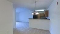 This single-story space showcases an open-concept layout that seamlessly connects the kitchen and living area. The vaulted ceiling adds a sense of volume and airiness to the interior, creating a welcoming and expansive atmosphere throughout.

The kitchen is defined by a peninsula with a dark countertop, providing both a workspace and a natural divider between the cooking and living zones. Medium-toned wood cabinetry lines the kitchen walls, offering ample storage, while a black refrigerator anchors the space. An under-cabinet fluorescent light fixture provides functional task lighting over the work area. A decorative multi-arm chandelier with frosted glass shades hangs above the peninsula, adding a traditional design touch.

The adjoining living area is finished with neutral-toned carpeting that extends throughout, contributing to a soft, comfortable feel underfoot. The walls are painted a clean, crisp white, reflecting light and enhancing the open character of the space. The layout flows naturally, making the combined kitchen and living area practical for everyday use and entertaining alike.