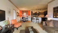 This single-floor space showcases an open-concept layout that integrates the living, dining, and kitchen areas into one cohesive environment. The most striking design element is the bold orange accent wall in the dining area, which anchors the space and adds dramatic warmth. Framed wall art and decorative panels complement the vibrant backdrop, enhancing the visual appeal.

The dining area features a dark wood table with cross-back chairs, set atop a warm-toned area rug. Adjacent to it, the kitchen island offers additional bar-style seating with two tall stools, providing a casual dining option. The kitchen itself is equipped with dark espresso cabinetry, a tile backsplash with contrasting geometric accents, a microwave, and ample counter space illuminated by pendant lighting suspended from the ceiling.

The living area is furnished with a gray sofa, an orange accent chair, and a dark wood coffee table with decorative inlay panels, all arranged over a patterned area rug. Track lighting runs along the ceiling, supplementing the pendant fixtures to ensure the entire space is well-lit. Neutral carpet flooring throughout ties the zones together, creating a warm and inviting atmosphere.