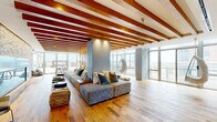 The 8th Floor lounge is a thoughtfully designed communal space that combines natural materials, contemporary furnishings, and panoramic views to create a sophisticated and inviting atmosphere. Rich hardwood flooring runs throughout, complementing the striking exposed wood beam ceiling that defines the main lounge area and adds warmth and architectural character to the space.

The primary lounge zone features an extended, modular sectional sofa with layered throw pillows, flanked by woven accent ottomans and low-profile side tables. Suspended egg-shaped hanging chairs add a playful, design-forward element, while vertical rope and wood dividers provide subtle spatial definition without obstructing sightlines. Floor-to-ceiling windows wrap the perimeter, flooding the interior with natural light and offering expansive city and skyline views.

At one end, a dramatic stone-clad fireplace serves as a focal point, framed by built-in shelving niches and a sleek glass-front firebox. Adjacent to the main lounge, a well-appointed wet bar features stone countertops, a stainless steel sink, and integrated appliances beneath a slatted wood ceiling, ideal for entertaining. Smaller bistro seating near the windows provides intimate spots to take in the surrounding urban views.