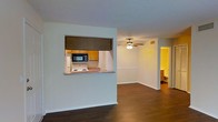This single-floor residence offers a functional and open layout designed for comfortable everyday living. The living area is a defining feature of the space, highlighted by dark hardwood flooring that extends throughout and a ceiling fan that provides both style and airflow. Clean white walls and recessed lighting contribute to a bright, airy atmosphere.

The kitchen is accessed through a distinctive pass-through window framed by a dark header, creating a connected yet defined cooking space. Warm honey-toned cabinetry lines the kitchen walls, complemented by a white electric range, an undermount sink, and a black microwave. The pass-through counter provides a convenient surface that bridges the kitchen and living areas, enhancing the sense of openness and flow.

A hallway visible beyond the living room leads to additional areas of the home, with a warmly lit room — appearing to be a bathroom — visible in the background, featuring yellow-toned lighting. The textured ceiling adds a classic architectural touch consistent throughout the space.

Overall, the layout is practical and well-connected, with each area transitioning naturally into the next. The combination of hardwood floors, warm cabinetry tones, and open sightlines gives the space a welcoming and livable character.