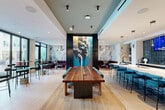Located at 3310 S Tryon St in Charlotte, NC, this single-floor amenity lounge offers a thoughtfully curated communal space spanning a generous footprint. The design aesthetic blends contemporary sophistication with bold artistic expression, highlighted by a striking large-scale mural of a figure rendered in teal, black, and gold that anchors the central gathering area.

Light wood-tone flooring runs throughout, contrasting elegantly with dark architectural accents and sleek black-framed glass partitions that define distinct zones while maintaining an open, connected feel. A solid live-edge walnut communal table with teal upholstered seating serves as the centerpiece of the main lounge area, complemented by a white marble-style bar counter with bar-height seating along one wall.

Multiple seating areas feature plush velvet furnishings in deep reds and burgundies, offering both intimate and social configurations. Vertical wood slat dividers create semi-private nooks filled with natural light from floor-to-ceiling windows that frame outdoor views of the surrounding Charlotte streetscape.

Glass-enclosed private rooms provide more secluded settings, one featuring a curved sectional sofa, ambient lighting, and an entertainment screen. A built-in wet bar with teal tile backsplash and open shelving adds functional convenience. Abstract wall murals and artistic wallcoverings throughout reinforce the space's gallery-like ambiance.