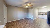 Located at 100 Horizon Ct NW in Concord, North Carolina, this single-floor unit offers a practical and efficient layout comprising a living area and a full bathroom. The main living space is finished with warm-toned wood-look plank flooring that runs throughout, providing a cohesive and clean aesthetic. Neutral gray walls complement the light-colored trim and white baseboards, creating a bright, understated interior. A ceiling fan with an integrated light fixture is centrally mounted above, offering both air circulation and ambient lighting. A wall-mounted window air conditioning unit provides dedicated climate control, while a window fitted with horizontal blinds allows for natural light and outside views. The layout also reveals a compact kitchen area adjacent to the main room, visible through the entryway, featuring white cabinetry. The bathroom is neatly appointed with a vanity featuring a dark granite-style countertop, white cabinetry below, and grab bars installed along the walls for added accessibility and safety. A textured ceiling throughout the unit adds a classic finishing detail. The front entry door opens directly into the living space, making the layout feel open and straightforward. Overall, the unit presents a tidy, functional living environment suited for single occupancy.