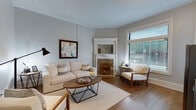 This single-floor residence blends classic character with modern updates throughout its well-appointed layout. The living room serves as the heart of the home, showcasing rich hardwood floors in a herringbone pattern, soft neutral walls, and abundant natural light streaming through large windows fitted with horizontal blinds. A standout decorative corner fireplace with ornate white millwork, a mirrored mantel, and detailed tile surround lends timeless charm to the space. The kitchen is thoughtfully designed with grey shaker-style cabinetry, white subway tile backsplash, stainless steel appliances including a gas range, microwave, dishwasher, and refrigerator, and a clean white countertop with an undermount sink — all combining functionality with a contemporary aesthetic. The bedrooms offer comfortable, carpeted retreats with ample closet space, including a louvered double-door closet, and windows that provide natural light. A dedicated desk area in one bedroom adds practical versatility. The bathroom is crisp and modern, featuring a grey vanity with white countertop, a large mirror, and a fully tiled walk-in shower. Throughout the residence, a cohesive grey-toned palette and carefully chosen finishes create a polished, inviting atmosphere.