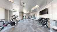 Located on Maple Street in Fort Collins, Colorado, this fitness center offers a comprehensive workout environment within a efficiently organized, single-floor layout. The space features a modern design with a drop-panel ceiling, recessed lighting, and neutral-toned carpet tiles laid in a grid pattern throughout.

The cardio section is well-stocked with multiple treadmills, elliptical trainers, upright stationary bikes, and a spin bike, several of which are equipped with personal display screens. Large windows along the perimeter flood the space with natural light while offering views of the surrounding outdoors, creating an open and energizing atmosphere.

The strength training area includes a power rack with an adjustable bench, a barbell, and a full dumbbell rack accommodating a wide range of weights. A wall-mounted flat-screen TV is positioned for visibility across the floor. Full-length mirrors line one wall, visually expanding the space and supporting proper form during exercise.

Additional amenities include wall-mounted yoga mat storage with hooks, a hand sanitizer station, and a glass-panel entry door connecting to an adjacent common area. The overall design is clean, bright, and functional, providing users with a complete fitness facility suitable for both cardio and strength-based training.