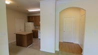 This single-floor residence offers a practical and well-connected layout. The kitchen is equipped with warm wood cabinetry, a white refrigerator, and a white range, all set atop light tile flooring. A peninsula counter extends into the space, providing additional workspace and a natural transition into the adjoining living area, which is finished with soft carpet flooring. A standout architectural feature is the decorative arched entryway framed by a column with crown molding detail, adding a classic, refined touch to the interior. This arch leads into a hallway where a paneled door provides access to an additional room. The neutral, light-toned walls throughout keep the space feeling open and cohesive. Crown molding accents the column, contributing to the overall traditional character of the home. The layout is straightforward and efficient, with the kitchen, living, and connecting hallway areas flowing naturally into one another, making the space feel accessible and easy to navigate.