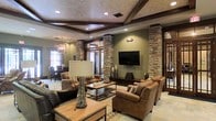 This single-floor space showcases a striking blend of rustic and refined design elements that create a warm, inviting atmosphere throughout. The living area serves as the focal point, anchored by bold stacked stone columns that rise from floor to ceiling, adding dramatic architectural character and a natural, earthy texture to the space. Dark wood beams frame a coffered ceiling with recessed lighting, contributing depth and a sense of grandeur overhead.

The seating arrangement features generously sized leather sofas and armchairs in rich cognac tones, gathered around a wood-and-metal coffee table atop a patterned area rug. A wall-mounted flat-screen television is positioned between the stone columns, creating a natural media wall. Sage green walls complement the stone and wood tones throughout, lending a cohesive, nature-inspired palette.

Visible in the background, a dining area with traditional furnishings and a chandelier adds functional elegance. Large windows allow natural light to filter into both areas. The overall ambiance is sophisticated yet comfortable, evoking a high-end lodge aesthetic well-suited for both relaxed gatherings and everyday living.