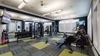 This fitness center is designed to offer a comprehensive workout environment within a single-level layout. The space is defined by bold navy blue walls accented with crisp white crown molding and trim, creating a polished, energetic atmosphere. Large windows with transom panels line multiple walls, allowing generous natural light to fill the room while maintaining privacy through adjustable horizontal blinds.

The flooring consists of modular carpet tiles in a patchwork pattern of chartreuse green and striped blue-gray, adding visual interest and defining workout zones throughout the space. Overhead, flush-mounted linear fluorescent fixtures and a ceiling fan provide consistent, even illumination.

The cardio zone features elliptical trainers, a recumbent bike, and a treadmill, each equipped with individual display screens. A Bodycraft rowing machine occupies open floor space in the center of the room. The strength-training area includes a multi-station cable machine, a weight bench, and a rack of dumbbells. Floor-to-ceiling mirrors along one wall visually expand the space and assist exercisers with form.

Near the entrance, a built-in shelving unit with lower cabinetry provides storage, while wall-mounted flat-screen televisions are positioned for visibility throughout the room. The overall design balances functionality with a modern, inviting aesthetic.