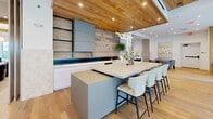 Located at Patriots Point in Mount Pleasant, South Carolina, this thoughtfully designed single-floor space offers a generous, open-concept layout ideal for gathering and entertaining. Wide-plank light oak hardwood flooring runs throughout, unifying the various living zones with a warm, cohesive aesthetic.

The centerpiece of the space is a stunning designer kitchen featuring a large island with seating for several, sleek slate-blue cabinetry, a wood-plank accent wall, and a sculptural pendant light fixture. A deep navy backsplash and open shelving add depth and character to the culinary area.

The main living room is anchored by a dramatic wood-paneled feature wall with a mounted TV, complemented by floor-to-ceiling glass doors that invite natural light and provide outdoor access. A second, more intimate living area includes a linear fireplace, a glass sliding partition, and refined furnishings. A third lounge area features an exposed brick accent wall with a marble floating shelf and mounted display.

The formal dining room is distinguished by an exposed wood beam ceiling, a statement chandelier, and a long communal table surrounded by upholstered chairs. Built-in shelving lines the walls, adding both function and visual interest.

Overhead, wood-clad ceiling panels with recessed lighting add warmth and architectural distinction throughout, creating a refined yet welcoming atmosphere.