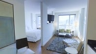 This single-floor open-concept studio is designed with a clean, modern aesthetic, characterized by crisp white walls and warm hardwood flooring throughout. A partial dividing wall creates a natural separation between the sleeping and living areas while maintaining an open, airy feel. The sleeping area accommodates a full-sized bed dressed in white linens, complemented by a wall-mounted flat-screen television positioned at the junction between the two spaces, making it viewable from both areas. The living area is furnished with a contemporary grey sofa and matching chairs accented by green throw pillows, anchored by a textured area rug and a sleek glass coffee table. A tufted white ottoman adds additional seating flexibility. Large windows flood the space with abundant natural light and frame expansive city skyline views, contributing to the unit's bright, elevated atmosphere. The overall design is minimalist and cohesive, with neutral tones and tasteful modern furnishings creating a comfortable and functional urban living environment.
