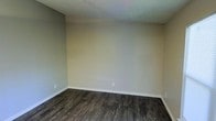 This single-floor space presents a straightforward and functional interior with a clean, move-in-ready aesthetic. The flooring throughout features rich, dark hardwood-style planks with a weathered wood grain pattern, adding depth and character to the space. The walls are finished in a warm neutral tone, creating a calm and adaptable backdrop that pairs well with a variety of décor styles. White baseboards provide a crisp contrast against both the flooring and the walls, lending a tidy, well-maintained appearance. The ceiling features a classic textured popcorn finish. Electrical outlets are positioned at standard heights along the walls, reflecting a practical layout. The open, unobstructed nature of the room provides flexibility for a range of uses, from a living area to a bedroom or home office. The overall ambiance is understated and neutral, making it an ideal canvas for customization. The combination of dark flooring and light walls creates a balanced contrast that gives the space a grounded, cohesive feel throughout.
