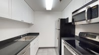 This space showcases a well-organized galley-style kitchen designed for functional, everyday living. The kitchen follows a classic parallel layout, with cabinetry and countertops running along both sides to maximize efficiency and workflow. White shaker-style cabinets extend from floor to ceiling on the left wall, providing ample storage space, while dark countertops create a striking contrast that anchors the room's clean aesthetic. A stainless steel sink is positioned along the left side, accompanied by additional lower cabinetry and what appears to be a dishwasher beneath the counter. On the right side, a full suite of stainless steel appliances lines the wall, including a refrigerator, an over-the-range microwave, and an electric range with a smooth cooktop — all contributing to a cohesive, modern look. The light tile flooring and bright white walls reflect the overhead fluorescent lighting, keeping the space feeling open and airy despite its compact form. The overall ambiance is crisp, neutral, and functional, making it a practical kitchen well-suited for everyday cooking needs.