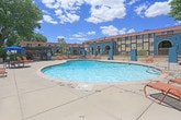 Located at 4217 Louisiana Blvd NE in Albuquerque, New Mexico, this outdoor amenity area showcases a vibrant Southwestern architectural style defined by bold teal stucco walls, terracotta red-tiled rooflines, and arched detailing throughout the surrounding structures. At the heart of the space is a generously sized freeform swimming pool with crystal-clear blue water, framed by a broad concrete pool deck furnished with terracotta-colored lounge chairs and a shaded umbrella table — ideal for relaxing under Albuquerque's abundant sunshine. Mature shade trees are positioned throughout the grounds, providing natural canopy and softening the open-sky setting. A gated pergola area, constructed with dark-stained wood beams and supported by substantial teal stucco columns, offers a covered outdoor lounge space featuring a stone fire pit surrounded by cushioned seating — creating an inviting spot for evening gatherings. The entrance to this area is accessed through a wrought iron gate set within brick-paved pathways, adding a sense of enclosure and privacy. The overall ambiance balances the warmth of New Mexican design traditions with practical outdoor living features, all set against the backdrop of New Mexico's characteristic wide-open blue skies.
