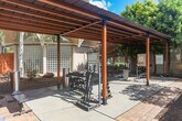 Located in Santa Clara, California, this thoughtfully designed outdoor space combines fitness functionality with attractive landscaping and architectural character. At its core is a spacious covered patio structure, built with bold steel posts and warm wood-toned beams supporting a corrugated metal roof. This pergola-style shelter provides shade over a concrete fitness area equipped with dumbbells, kettlebells, a weight rack, and a workout bench — offering a well-appointed open-air gym experience.

A standout architectural feature is the large glass block wall, framed beneath a curved canopy with rounded white columns, which diffuses natural light and adds a distinctive mid-century modern touch to the space. The surrounding landscape is carefully curated, with horizontal cedar-style privacy fencing enclosing the area, mature birch trees, Japanese maples displaying seasonal color, ornamental shrubs, and decorative boulders nestled into mulched garden beds. A paved walkway connects the various zones, blending functionality with aesthetic appeal. The open sky above and abundant greenery create a bright, welcoming environment that balances active use with natural tranquility.