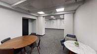 Located at 888 N Main St in Santa Ana, California, this single-floor commercial space offers a clean, professional environment designed for both collaborative and private use. The layout is thoughtfully organized, combining open lounge areas with a dedicated meeting room to accommodate a variety of functional needs.

The open areas feature patterned gray carpeting and crisp white walls, creating a neutral, distraction-free backdrop. Recessed panel lighting in the drop ceiling provides consistent, even illumination throughout the space. Multiple seating arrangements are distributed across the open zones, including mid-century-style navy blue armchairs paired with small round side tables — ideal for informal gatherings or breakout discussions.

A private meeting room anchors one section of the space, furnished with a circular wood-topped conference table surrounded by upholstered black chairs. The walls of this room are adorned with a curated selection of abstract artwork in cool blue and neutral tones, adding a refined aesthetic touch.

Structural columns and defined wall recesses lend architectural character to the otherwise open floor plan, while dark-framed access doors provide clear transitions between zones. The overall ambiance is polished and understated, making this space well-suited for professional, office, or event-oriented use.