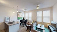 The Bay Front is a well-appointed 1-bedroom, 1-bathroom residence offering a thoughtfully designed single-level layout. The open-concept living and dining area is bright and welcoming, with large windows dressed in white horizontal blinds that allow generous natural light to fill the space. Wood-look flooring runs throughout the main living areas, adding warmth and continuity to the design.

The kitchen is a functional centerpiece, featuring granite countertops, a large island with an undermount sink, stainless steel appliances including a refrigerator, range, and microwave, and contemporary wood-toned cabinetry. Pendant lighting above the island and track lighting overhead enhance both style and practicality. The adjacent dining area comfortably accommodates a table and chairs, making it ideal for everyday meals or entertaining.

The living room offers ample space for seating and entertainment, with a ceiling fan overhead and multiple windows providing cross-ventilation and natural brightness.

The bedroom is a calm, carpeted retreat with a ceiling fan, warm ambient lighting, and room for a full dresser and nightstands. Framed artwork adds a polished finishing touch to the space.

The bathroom includes a full soaking tub with subway tile surround, a granite vanity countertop, and clean, neutral finishes throughout.
