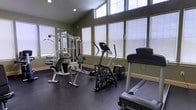 This fitness center offers a dedicated space for a comprehensive workout experience. The room is defined by its vaulted ceiling, which lends an open, airy feel, while a row of large windows along the upper and lower walls invites abundant natural light, creating a bright and motivating environment. The walls are finished in a deep olive green, giving the space a focused and energizing ambiance, complemented by a dark, durable rubber flooring ideal for high-impact exercise. The facility is equipped with a multi-station cable and weight machine, providing a versatile range of strength training exercises. Cardio options include an elliptical cross-trainer and a treadmill, accommodating a variety of fitness routines. A wall-mounted mirror enhances the sense of space while allowing users to monitor their form during workouts. The room's layout is thoughtfully arranged to allow comfortable movement between equipment. Roller shades on the windows offer adjustable light and privacy control. Overall, this fitness center combines practical functionality with a clean, modern design, making it a well-rounded amenity for daily exercise.