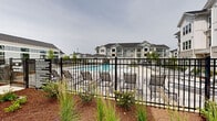 Located at 601 Cambrian Ridge Trail in Wake Forest, NC, this three-story residential community showcases a contemporary craftsman-inspired architectural style, combining board-and-batten siding, brick accents, and dark pitched rooflines across multiple well-designed buildings. The exterior grounds are thoughtfully landscaped with manicured lawns, young trees, mulched garden beds, and curving concrete walkways that connect the community's many amenity areas.

At the heart of the community lies a resort-style swimming pool surrounded by a spacious concrete deck lined with lounge chairs, offering ample space for relaxation. Adjacent to the pool, a pergola-covered terrace provides shaded seating with stylish outdoor furniture, while a separate turf lawn area features additional chaise lounges beneath a slatted overhead structure. Restroom facilities are conveniently located poolside.

A dedicated bocce ball court adds a recreational focal point to the outdoor amenity corridor, framed by string lights and a white pergola structure with comfortable seating nearby. Green lawn areas throughout the community offer open gathering spaces, and covered pergola stations with Adirondack-style chairs provide inviting spots for residents to relax outdoors. Resident balconies are visible across all three floors, and ample parking with landscaped surroundings completes this well-appointed community.
