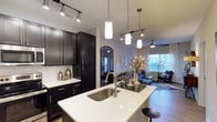 This single-floor, one-bedroom, one-bathroom residence showcases a thoughtfully designed open-concept layout that connects the kitchen and living areas into one cohesive space. The kitchen is outfitted with dark espresso cabinetry, crisp white quartz countertops, and a subway tile backsplash that adds a clean, contemporary contrast. A large kitchen island with an undermount stainless steel sink serves as a functional centerpiece, complemented by stylish pendant lighting that hangs overhead. Track lighting along the ceiling provides additional illumination throughout the kitchen. An arched decorative mirror accent adds architectural character and visual depth to the transitional zone between the kitchen and living area. The living space is bright and welcoming, with natural light streaming through large windows dressed with sheer curtains. Wood-style flooring runs throughout, lending warmth and continuity across the open floor plan. A ceiling fan in the living area adds both comfort and style. The overall ambiance is modern and refined, balancing dark cabinetry tones with light countertops and neutral walls for an inviting, well-appointed feel.