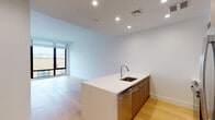 Located at 15 Bond Street in Great Neck, NY, this modern single-floor residence showcases a clean, contemporary design throughout. The open-concept living and kitchen area is anchored by a sleek kitchen island with a white quartz countertop and an undermount sink, complemented by warm wood-toned cabinetry that extends to the upper walls. Stainless steel appliances — including a full-size refrigerator, range, and microwave — provide a polished, functional finish. Light-toned hardwood flooring runs seamlessly throughout the main living areas, enhancing the sense of openness and warmth. Large floor-to-ceiling windows allow generous natural light to fill the space while offering views of the surrounding neighborhood. The living area is spacious and versatile, with crisp white walls and recessed lighting that contribute to the unit's bright, airy ambiance. The bathroom features a modern aesthetic with dark slate-toned tile flooring, a light wood vanity with an undermount sink, a large framed mirror, and a walk-in shower, creating a clean and refined feel. Overall, the residence presents a cohesive, minimalist design that balances functionality with contemporary style, making it an ideal space in the heart of Great Neck.