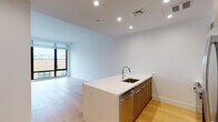 Located at 15 Bond Street in Great Neck, NY, this modern single-floor residence showcases a clean, contemporary design throughout. The open-concept living and kitchen area is anchored by a sleek kitchen island with a white quartz countertop and an undermount sink, complemented by warm wood-toned cabinetry that extends to the upper walls. Stainless steel appliances — including a full-size refrigerator, range, and microwave — provide a polished, functional finish. Light-toned hardwood flooring runs seamlessly throughout the main living areas, enhancing the sense of openness and warmth. Large floor-to-ceiling windows allow generous natural light to fill the space while offering views of the surrounding neighborhood. The living area is spacious and versatile, with crisp white walls and recessed lighting that contribute to the unit's bright, airy ambiance. The bathroom features a modern aesthetic with dark slate-toned tile flooring, a light wood vanity with an undermount sink, a large framed mirror, and a walk-in shower, creating a clean and refined feel. Overall, the residence presents a cohesive, minimalist design that balances functionality with contemporary style, making it an ideal space in the heart of Great Neck.