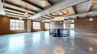This expansive single-floor space showcases a striking industrial aesthetic, characterized by a thoughtful blend of raw and refined architectural elements. The open layout offers a generous, column-free expanse that is highly adaptable for a variety of uses, from event hosting to creative office or retail configurations.

Exposed brick walls line the perimeter, lending warmth and texture to the space, while the polished concrete floors reflect abundant natural light that pours in through rows of large, multi-pane industrial windows. The coffered concrete ceiling, supported by heavy timber beams, adds structural character and visual depth overhead. A series of pendant industrial-style light fixtures hang throughout, complementing the building's heritage character.

A central curved spiral staircase with a sleek black metal railing serves as a dramatic architectural focal point, drawing the eye and providing vertical connectivity. A pair of stainless steel elevators, framed by exposed brick, offer accessible entry to the floor.

The windows along multiple walls frame views of the surrounding urban landscape, providing a strong connection to the exterior environment. Exposed mechanical systems on the ceiling contribute to the authentic industrial feel. This space combines historic building character with modern utility, offering a versatile and visually compelling environment.