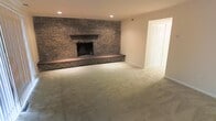 This two-floor space offers a well-structured layout that balances open living areas with practical design. The standout feature of the main living area is a full-height exposed brick fireplace wall, lending the room a warm, rustic character. The fireplace is framed by a classic wood mantel and raised brick hearth, serving as a natural focal point for the room. Soft, neutral-toned carpet flows throughout the space, contributing to a cozy and inviting ambiance. Recessed lighting in the ceiling provides clean, even illumination without disrupting the room's understated aesthetic. Smooth, light-painted walls complement the textural contrast of the brick, creating a balanced visual composition. A built-in closet with louvered doors offers discreet storage within the living area, adding to the space's functionality. The overall design reflects a traditional sensibility with durable, low-maintenance finishes. The generous proportions of the living room allow for flexible furniture arrangements to suit a variety of uses. Across its two floors, the space provides ample room for both daily living and entertaining, making it a versatile and well-appointed property.