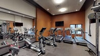 This fitness center is thoughtfully designed to support a full range of workout activities within a compact, efficiently organized space. The room features warm terracotta-toned accent walls paired with neutral gray upper walls and a drop-panel ceiling outfitted with recessed and flush-mount lighting, creating a focused and energizing atmosphere. Durable speckled rubber flooring covers the entire space, providing traction and cushioning underfoot.

The cardio zone is prominently positioned along one wall and includes multiple Life Fitness treadmills and elliptical cross-trainers, all facing a wall-mounted flat-screen television for entertainment during workouts. A window provides a modest view to the exterior, adding a sense of openness. Large mirrors along adjoining walls visually expand the space and allow users to monitor their form throughout exercises.

The strength training area features a multi-cable functional trainer system suitable for a wide variety of resistance exercises, complemented by a dumbbell rack, an adjustable workout bench, a balance dome, and a stretch board. A workout reference poster is mounted on the wall for guidance. Additional amenities include a water cooler station, towel storage, and a wall clock, ensuring users have everything needed for a convenient and complete fitness session.