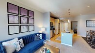 This single-floor residence showcases an open-concept design that connects the living area and kitchen in a fluid, functional layout. The living space features a bold royal blue sofa as a centerpiece, set against light gray walls adorned with a gallery arrangement of framed artwork in soft purple and white tones. Recessed lighting and a classic table lamp provide warm, layered illumination throughout.

The carpeted living area transitions smoothly into hardwood flooring that extends through the kitchen zone. The kitchen is well-appointed with light maple cabinetry, stainless steel appliances including a refrigerator, microwave, and range, and a peninsula countertop with an undermount sink that doubles as a casual breakfast bar. Pendant lights hang above the peninsula, adding a modern decorative touch.

The overall color palette — soft grays, warm wood tones, and white countertops — creates a clean, contemporary ambiance. The open sightlines between the living and kitchen areas enhance the sense of space and make the layout ideal for both everyday living and entertaining.