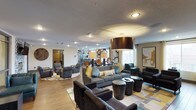 This single-floor space is a thoughtfully designed communal lounge that blends modern aesthetics with a warm, inviting atmosphere. The open-plan layout is anchored by rich wood-look flooring and defined by a series of distinct yet connected seating areas, creating a natural flow throughout the space. A large, oversized bronze drum pendant light serves as a striking focal point, complementing the warm tones of the décor and casting a soft, ambient glow across the room. Multiple seating arrangements feature plush dark charcoal sofas and armchairs, accented with mustard yellow and teal throw pillows that add pops of color against the neutral palette. Geometric patterned area rugs in blue, gold, and grey ground each seating cluster, lending structure to the open floor plan. In the background, a dining or café-style area is visible, illuminated by a cluster of pendant lights that add depth and visual interest. Large windows allow natural light to filter into the space, enhancing the bright and airy feel. Decorative wall art, sculptural table accents, and warm wood paneling details contribute to the polished, contemporary character of this well-appointed communal retreat.