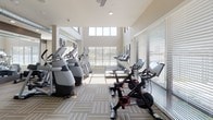 This single-level fitness center is designed to deliver a bright, motivating workout environment. The space is architecturally distinguished by its double-height ceilings in the main cardio zone, where oversized grid-pane windows stretch from mid-wall to the ceiling, flooding the floor with natural light and offering views of the surrounding landscape. Throughout the facility, neutral-toned patterned carpet tiles provide a cohesive, commercial-grade foundation underfoot.

The main cardio area is generously lined with treadmills, each equipped with individual monitor screens, positioned directly facing the large windows. Elliptical trainers and stair climbers from Precor are arranged alongside, maximizing the light-filled perimeter. Ceiling fans are installed throughout to maintain airflow and comfort.

A secondary zone transitions into a functional training area, featuring Peloton spin bikes, rowing machines, and a heavy tire for functional strength work. Exposed ductwork along the ceiling adds an industrial design element to this section. Floor-to-ceiling mirrors visually expand the space and allow users to monitor their form during exercise.

Natural light is abundant throughout, supplemented by recessed lighting and ceiling fans. The overall ambiance is clean, modern, and energizing, well-suited for a full range of fitness activities.