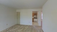 This single-floor residence offers a straightforward and functional layout designed for comfortable everyday living. The main living area is finished with wall-to-wall carpeting in a neutral tone, creating a warm and inviting atmosphere. Smooth white walls and a clean ceiling contribute to a bright, airy feel throughout the space. A louvered closet door provides convenient built-in storage within the living area, adding to the room's practicality. An open passageway connects the living space directly to the kitchen, promoting an easy flow between the two areas. The kitchen features warm-toned oak cabinetry that provides ample storage, paired with a white gas range and matching appliances. Light-colored flooring in the kitchen contrasts gently with the carpeted living space, visually defining each zone. A window in the adjacent area allows natural light to filter in, brightening the interior. The overall design is clean and neutral, offering a versatile canvas that can be easily personalized. The seamless connection between the living and kitchen areas makes this space well-suited for both relaxed daily routines and casual entertaining.