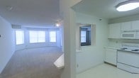 This single-floor residence offers a clean, open layout designed for comfortable everyday living. The living area is generously proportioned, featuring soft wall-to-wall carpeting that adds warmth underfoot and a track lighting fixture overhead that provides focused illumination throughout the space. Multiple windows line the walls of the living area, flooding the room with natural light and creating a bright, airy atmosphere.

A structural dividing wall with a pass-through window connects the living area to the kitchen, allowing for an open sightline between the two spaces while maintaining a sense of defined zones. The kitchen features light tile flooring and white cabinetry, giving it a clean and functional aesthetic that complements the neutral tones found throughout the residence.

The overall color palette is kept light and neutral, with white walls and ceilings that enhance the sense of openness and allow for versatile interior styling. The layout flows intuitively from one area to the next, making the space feel cohesive and easy to navigate. This residence is well-suited for those seeking a bright, move-in ready space with a straightforward, practical design.