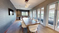Located at 1585 Anderson Rd in McLean, Virginia, this sophisticated amenity space seamlessly blends indoor comfort with outdoor living. The interior room is anchored by a large, solid-wood communal table surrounded by white contemporary chairs, making it ideal for meetings, dining, or social gatherings. A striking cluster pendant chandelier with globe lights serves as a dramatic focal point overhead, while textured accent walls — including a bold geometric-patterned wallcovering and a dark mosaic tile backsplash — add visual depth. A wall-mounted flat-screen TV, decorative wall art, and a wet bar with under-counter refrigeration round out the interior's polished, modern aesthetic. Light wood-style flooring flows throughout, complementing the neutral gray tones. Full-height glass French doors connect the interior to a covered outdoor terrace, where a slatted wood ceiling with sleek ceiling fans creates a resort-like ambiance. The terrace is furnished with modular lounge sofas and multiple dining sets, all oriented to take advantage of sweeping, unobstructed views of the surrounding treetop canopy. Large-format tile flooring and clean railings complete the upscale outdoor experience.