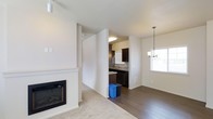 This single-floor 1-bedroom, 1-bathroom Hudson residence offers a well-planned open layout that connects the primary living spaces seamlessly. The living room serves as the focal point of the home, featuring a clean, white-framed fireplace that adds warmth and a contemporary touch. Plush carpeting in the living area provides comfort underfoot, while the adjoining dining and kitchen spaces transition to rich hardwood-style flooring, creating a clear and attractive visual distinction between zones.

The dining area benefits from natural light streaming through a window, complemented by an elegant chandelier that adds a refined detail to the space. A partial wall divides the living area from the kitchen, offering a sense of separation while maintaining an open feel. The kitchen is outfitted with dark cabinetry, mosaic tile backsplash, and dark appliances, creating a striking contrast against the light-toned walls. An additional window in the kitchen brings in further natural light. The overall color palette throughout the home is neutral and cohesive, with white walls and trim providing a bright, clean backdrop that makes the space feel welcoming and move-in ready.