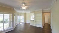 This single-floor space showcases a thoughtfully designed open-concept layout that seamlessly connects the living area to the kitchen. The living room is anchored by rich, dark hardwood flooring that extends throughout, complemented by warm beige walls and crisp white crown molding that frames the perimeter of the ceiling. A ceiling fan with an integrated light fixture is centrally positioned, providing both air circulation and ambient lighting.

A charming bay window area along one wall invites generous natural light, brightening the entire space and offering views of the surrounding exterior. The open shelving unit built into the wall provides practical and decorative display space, while a raised countertop pass-through connects the living area to the kitchen, maintaining an open flow while defining each zone. The kitchen, visible through the pass-through, features dark countertops contrasting with lighter cabinetry.

Crown molding details throughout and the clean, neutral color palette contribute to a polished, cohesive ambiance. The overall layout is well-proportioned, functional, and move-in ready.