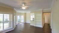 This single-floor space showcases a thoughtfully designed open-concept layout that seamlessly connects the living area to the kitchen. The living room is anchored by rich, dark hardwood flooring that extends throughout, complemented by warm beige walls and crisp white crown molding that frames the perimeter of the ceiling. A ceiling fan with an integrated light fixture is centrally positioned, providing both air circulation and ambient lighting.

A charming bay window area along one wall invites generous natural light, brightening the entire space and offering views of the surrounding exterior. The open shelving unit built into the wall provides practical and decorative display space, while a raised countertop pass-through connects the living area to the kitchen, maintaining an open flow while defining each zone. The kitchen, visible through the pass-through, features dark countertops contrasting with lighter cabinetry.

Crown molding details throughout and the clean, neutral color palette contribute to a polished, cohesive ambiance. The overall layout is well-proportioned, functional, and move-in ready.