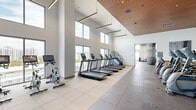Located at 1770 S Young Circle in Hollywood, Florida, this state-of-the-art fitness center spans a generous single floor divided into two dedicated exercise rooms. The facility is defined by its striking architectural design, featuring soaring double-height ceilings, warm wood-paneled ceiling accents, and floor-to-ceiling windows that flood the space with abundant natural light while offering panoramic outdoor views of the surrounding cityscape.

The cardio zone houses a full lineup of treadmills, ellipticals, stair climbers, rowing machines, and Matrix recumbent bikes, all positioned along the window-facing walls to maximize natural light and views. The strength and functional training area is equally well-appointed, featuring multi-station cable machines, weight racks stocked with barbells and dumbbells, resistance machines, medicine balls, stability balls, and kettlebells organized on dedicated storage racks.

Flooring transitions between large-format porcelain tile and resilient rubber gym flooring, delineating cardio and strength zones. Distinctive cylindrical metallic columns punctuate the open floor plan, adding an architectural flair. The facility also includes private rooms with textured tile accent walls, recessed and pendant lighting, and direct outdoor access, creating a bright, motivating, and fully comprehensive fitness environment.