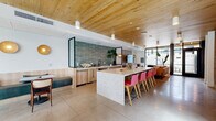 Located at 1501 Buena Vista Ave in Alameda, California, this single-floor communal space masterfully blends modern design with warm, natural materials. The open-plan layout seamlessly connects a well-appointed kitchen, casual dining area, and comfortable lounge zone, creating a versatile environment suited for both socializing and relaxation.

The kitchen area is anchored by a generously sized stone-topped island, finished in a speckled terrazzo-style material and surrounded by vibrant red bar stools with wood-frame legs. Light wood cabinetry lines the walls, complemented by a striking sage-green tile backsplash that adds a bold yet earthy contrast. A built-in sink and undercounter appliances complete the functional workspace.

Adjacent to the kitchen, a cozy dining nook features teal built-in bench seating paired with wooden chairs, lit by decorative gold disc wall sconces that lend a warm, ambient glow. The lounge area is anchored by a rich cognac leather sofa set against an exposed brick accent wall, adding an industrial character. Floor-to-ceiling black-framed glass doors and windows flood the space with natural light and offer direct views of and access to an outdoor area.

Throughout, natural wood plank ceilings tie the design together, creating a cohesive, resort-inspired aesthetic. Polished concrete floors provide a clean, durable foundation that unifies the entire space.