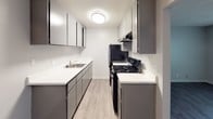 This 1-bedroom, 1-bathroom unit is thoughtfully designed across a single floor, offering a functional and comfortable living environment. The living area features light wood-style laminate flooring, soft neutral-toned walls, a textured ceiling, and a wall-mounted air conditioning unit, providing a clean and practical space for everyday living. A floor-standing heater unit is also present, ensuring year-round climate comfort.

The bedroom is generously sized, finished with plush blue-gray carpeting that adds warmth underfoot. A centered window frames views of mature trees outside, allowing natural light to fill the room and creating a calm, inviting atmosphere.

The galley-style kitchen is efficiently laid out with gray cabinetry on both sides, white quartz-style countertops, a single-basin sink, and black appliances including a gas range and refrigerator. Light wood-style flooring and flush ceiling lighting complete the kitchen's clean, modern aesthetic.

The bathroom features wood-look tile flooring, a white quartz vanity countertop, a full tub-shower combination, and brushed nickel fixtures and accessories. The neutral palette and simple design create a tidy, functional space.

Overall, the unit presents a cohesive, updated interior with consistent neutral tones and modern finishes throughout.